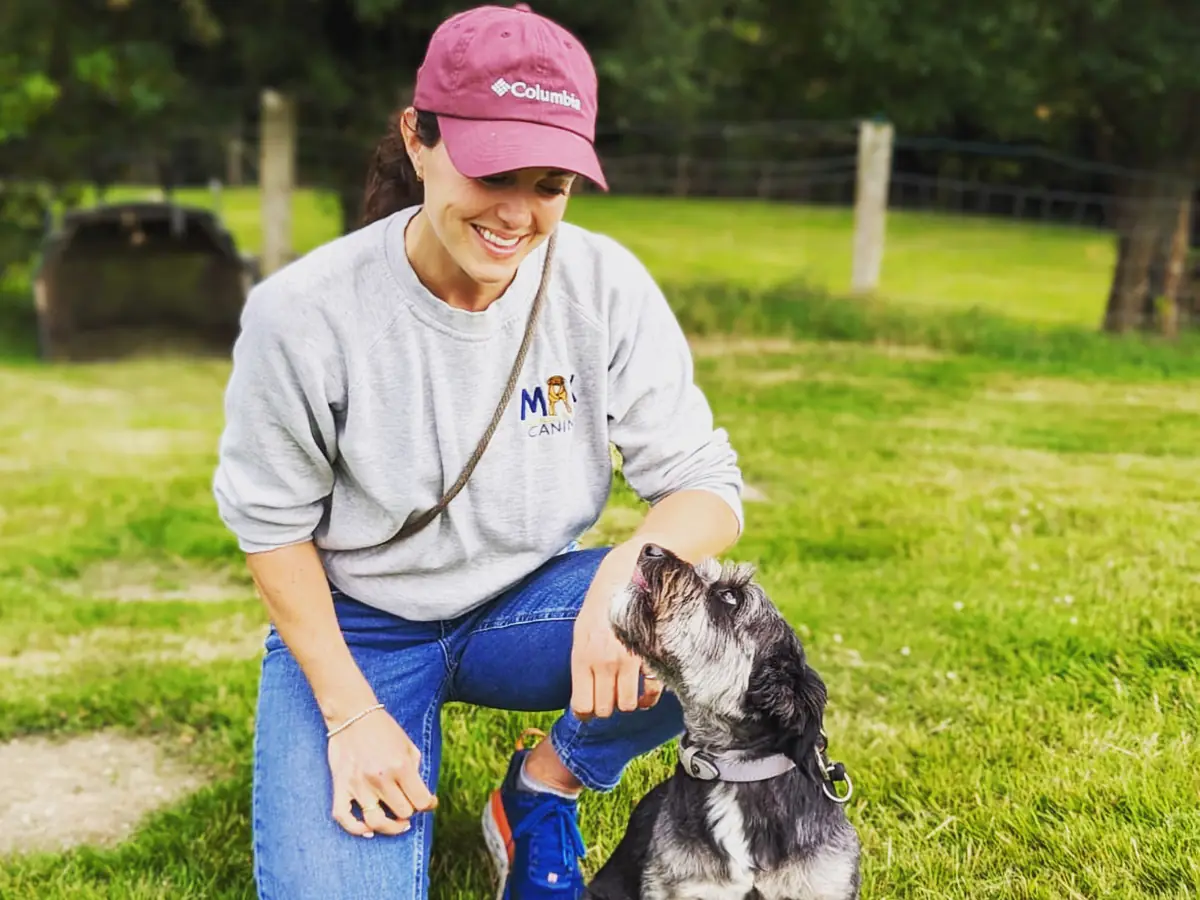 Emily kneeling on the grass in a Max Canine sweatshirt, smiling at a grey schnauzer cross looking up at her